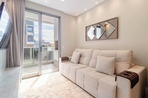A modern living room with a sofa, a sliding glass door, and a city view. - Luxury 2BR • 2 Min Walk to Beach • Rooftop Pool • BBQ • 2 Parking (Florianópolis)