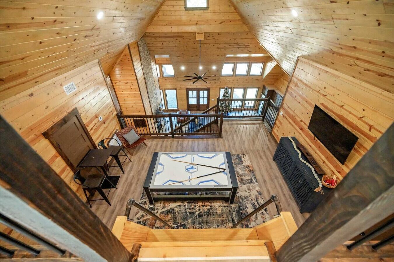 Interior de una cabaña de madera con una mesa central, una escalera y un televisor.