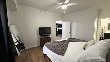 Habitación de hotel con cama, televisor en un soporte, ventilador de techo y vista a la cocina por una puerta abierta.