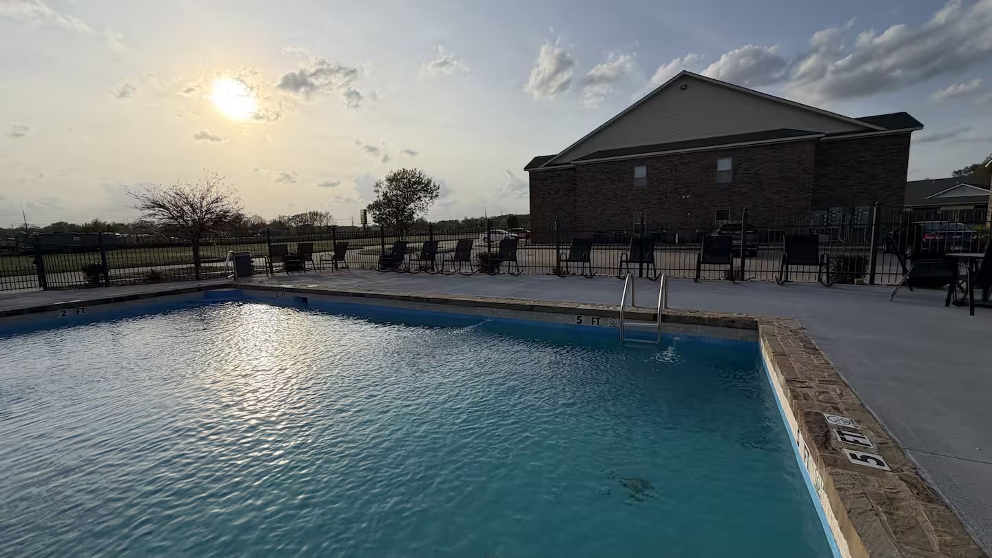 Una piscina con una escalera metálica, rodeada por una cerca, y un edificio con techo inclinado al fondo.