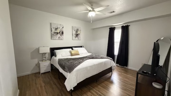 A bedroom with a bed, a nightstand, a desk, a ceiling fan, and artwork on the wall.