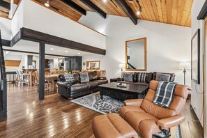 Ein geräumiges Wohnzimmer mit einer großen Couch, einem Couchtisch und einem Ledersessel. Es gibt einen Essbereich mit Tisch und Stühlen sowie eine Küche im Hintergrund.