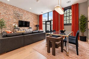 Ein modernes Wohnzimmer mit Backsteinwand, Flachbildfernseher, Glastisch mit Stühlen und einem großen Fenster mit roten Vorhängen.
