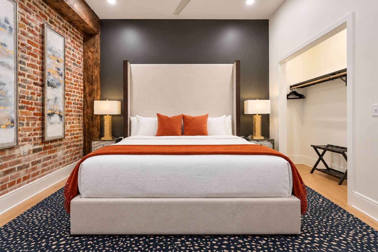 A hotel room with a large bed, two bedside tables with lamps, a brick wall with artwork, and a closet with a hanging rack.