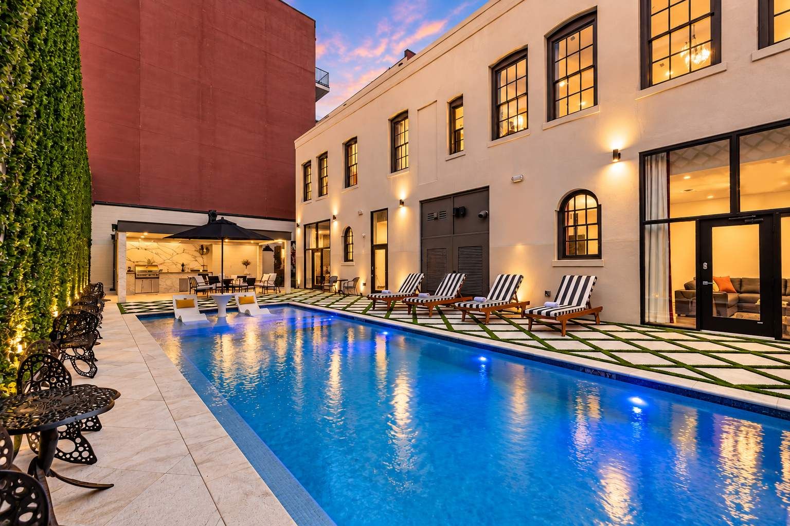 Una piscina con sillones de descanso y una zona de bar, rodeada de edificios con amplias ventanas y un muro rojo.