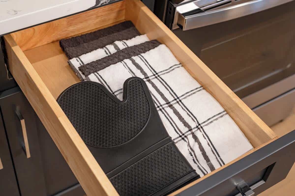 Un tiroir de cuisine contenant une spatule noire, un torchon à carreaux noirs et blancs, et un tapis noir texturé.