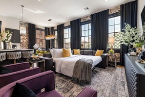 A modern hotel room with a large bed, a sitting area with a sofa and chairs, a bar area, and large windows with curtains.