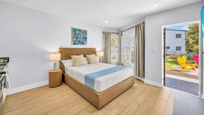A bedroom with a wooden bed, bedside lamps, a window with curtains, and a view of the outside. - Hawksbill 105 | Beach & Pool Studio | Walkable (Lauderdale-by-the-Sea)