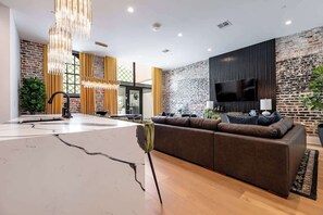 A modern living room with a large sectional sofa, a TV mounted on a brick wall, and a kitchen area with a marble countertop.