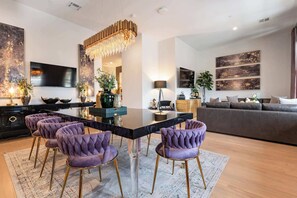 Un salon moderne avec un coin repas, comprenant une table à plateau en verre, des chaises à revêtement violet, un lustre et des œuvres d’art accrochées aux murs.