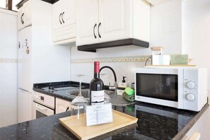 A kitchen with white cabinets, a microwave, a wine bottle, and a wooden cutting board with a menu. - Seafront Apt w/ Pool & Teide View, Near Dining (Punta del Hidalgo)