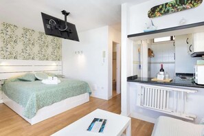 Ein Schlafzimmer mit Bett, Nachttisch, einem an der Wand befestigten Flachbildfernseher und einer Küchenzeile mit Arbeitsplatte und Herd.