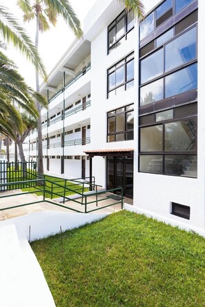 A modern multi-story building with a flat roof, large windows, and a covered outdoor area with a green lawn. - Seafront Apt w/ Pool & Teide View, Near Dining (Punta del Hidalgo)