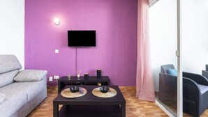 A living room with a purple wall, a flat-screen TV, a grey sofa, a black table with two woven placemats, and wicker furniture. - Apartamento Vistas Al Mar y Al Teide (Tacoronte)