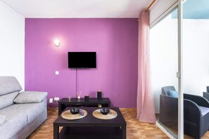 Ein Wohnzimmer mit einer violetten Wand, einem Flachbildfernseher, einem grauen Sofa, einem schwarzen Tisch mit zwei gewobenen Unterlagen und Korbmöbeln.