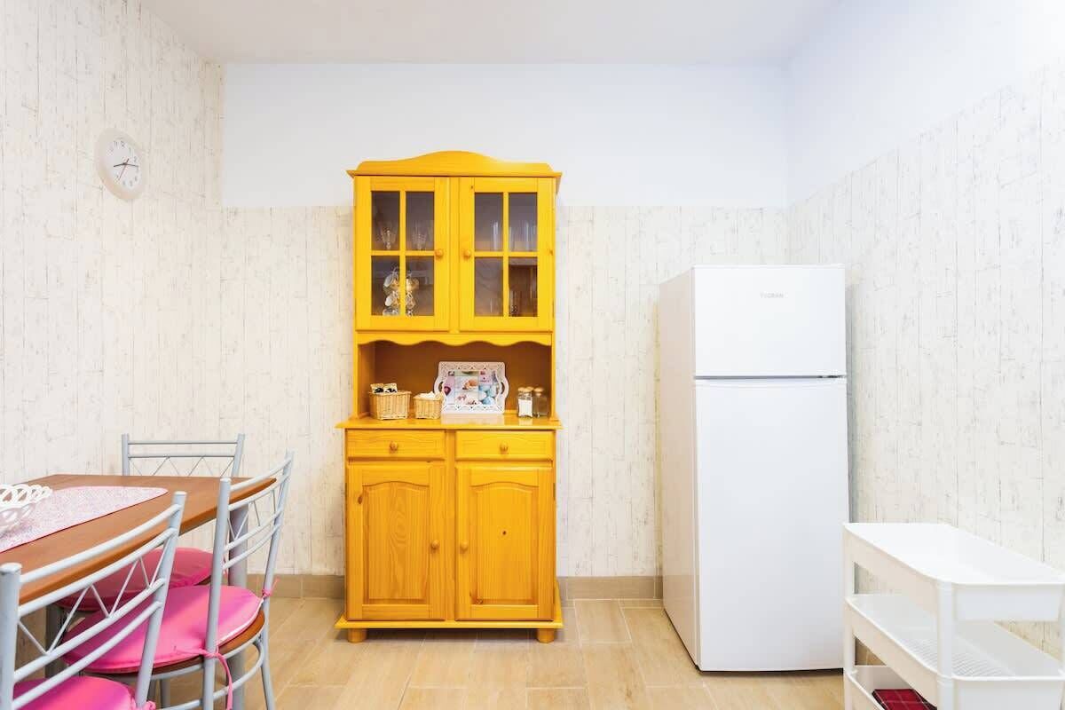 Une petite cuisine avec un réfrigérateur blanc, un meuble jaune aux portes en verre et une table en bois avec des chaises.