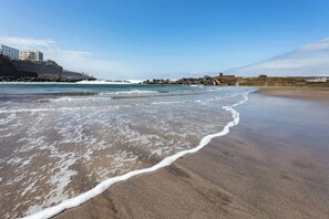 Ein Strand mit Wellen, einer felsigen Küstenlinie und Gebäuden im Hintergrund.