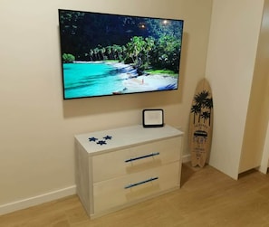 Ein an der Wand befestigter Fernseher, eine weiße Kommode mit Schubladen und ein Surfbrett, das an der Wand lehnt.