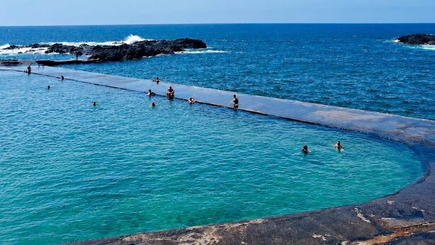 Een zwemgebied aan de kust met helderblauw water, rotsformaties en een lange pier die in zee uitloopt.