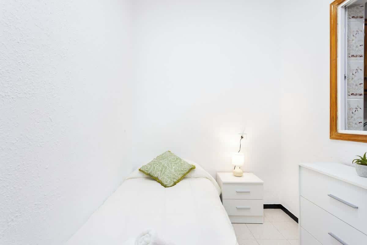 A small, clean room with a bed, a bedside table with a lamp, a dresser, and a framed picture on the wall.