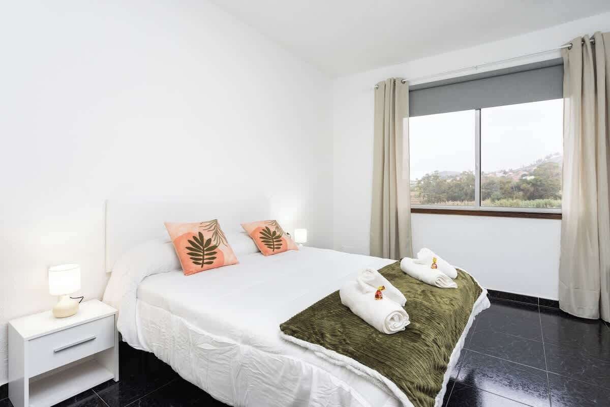A hotel room with a bed, a nightstand, a lamp, a window with curtains, and a view of greenery outside.