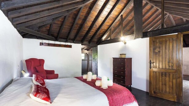 Um quarto com teto de madeira, uma cama com manta vermelha, uma cadeira vermelha, um guarda-roupa de madeira e uma porta de madeira.