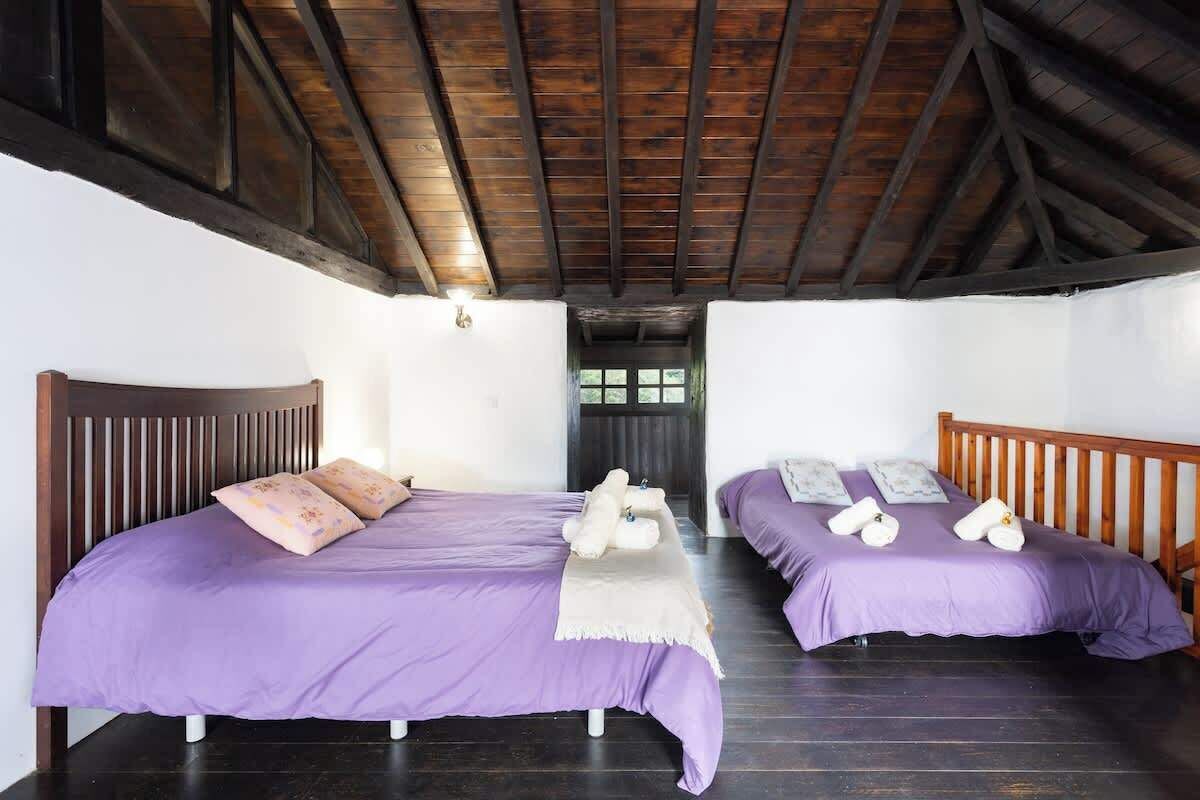 A room with two beds, wooden beams on the ceiling, and a window in the background.