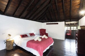 A bedroom with a wooden ceiling, a bed with white bedding and a red blanket, two red chairs, and a wooden dresser.