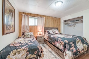 Un dormitorio con dos camas, cabeceros de madera, una mesita de noche, una lámpara, un cuadro enmarcado y un armario con perchas.