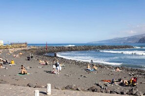 A beach with people sunbathing and walking along the shore, a rocky breakwater, and the ocean in the background. - Apartamento Terraza Martianez (Puerto de la Cruz)