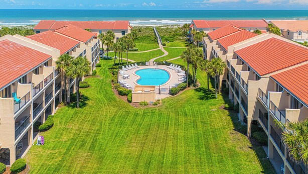 Un resort con una piscina centrale, diversi edifici con tetti in tegole rosse e vista sull'oceano.