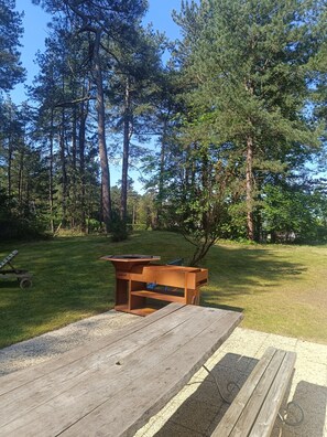 Eine Holzterrasse mit Grill, umgeben von einer Rasenfläche und hohen Bäumen.