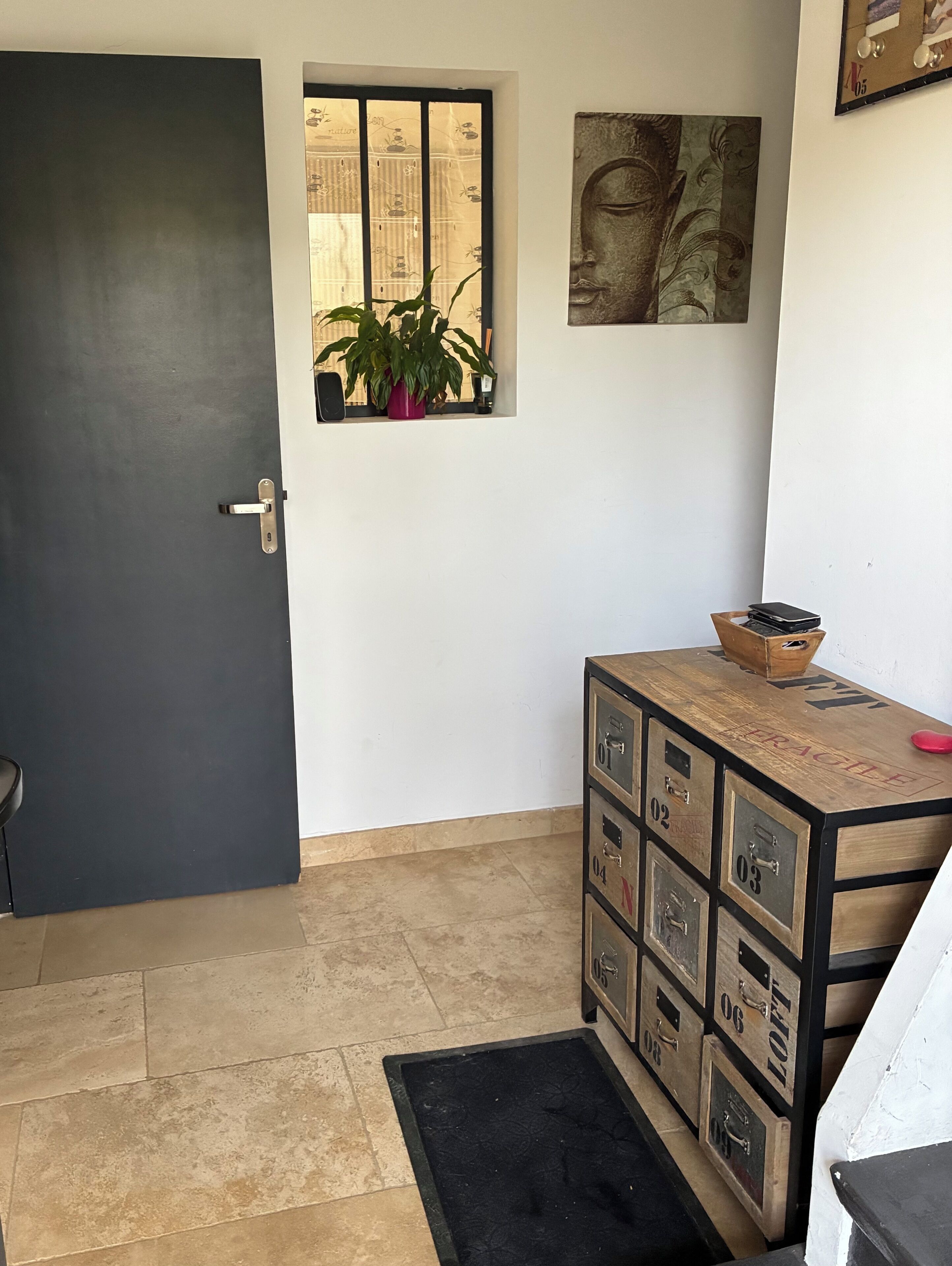 Una habitación con una puerta oscura, una ventana con una planta, un armario de madera con cajones y una obra de arte colgada en la pared.