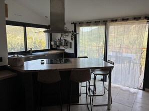 Eine moderne Küche mit einer großen Kochinsel, Barhocker, einem Fenster mit Blick auf Bäume und einem Edelstahl-Wasserkocher.