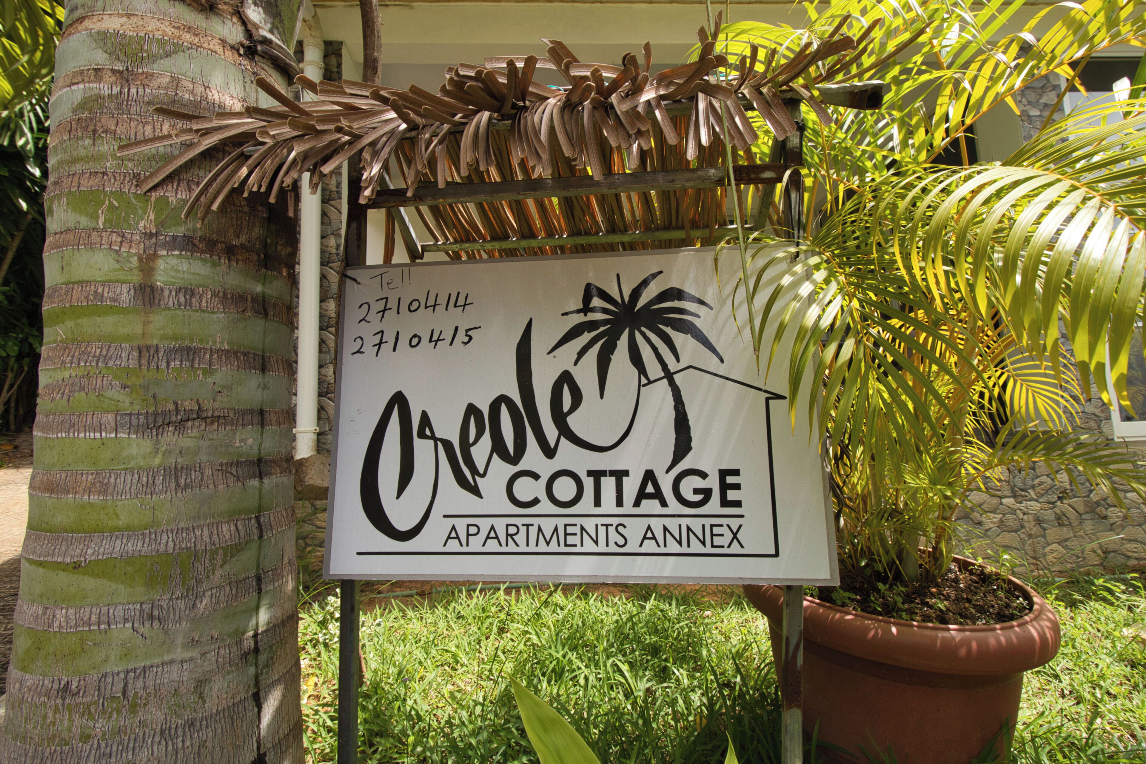 Une enseigne pour le complexe d’appartements Creole Cottage, avec un palmier et des plantes en pot.