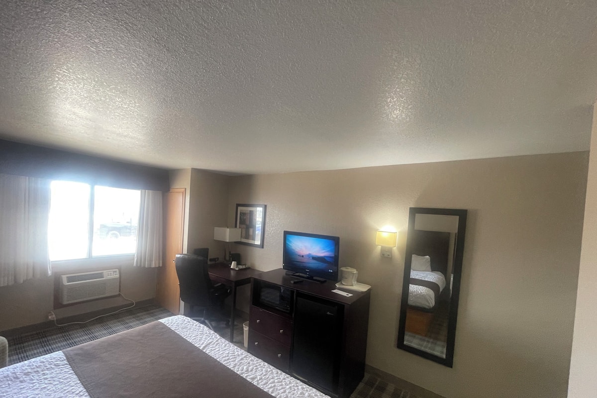 Habitación de hotel con cama, escritorio con computadora, televisor, espejo y ventana con cortinas.