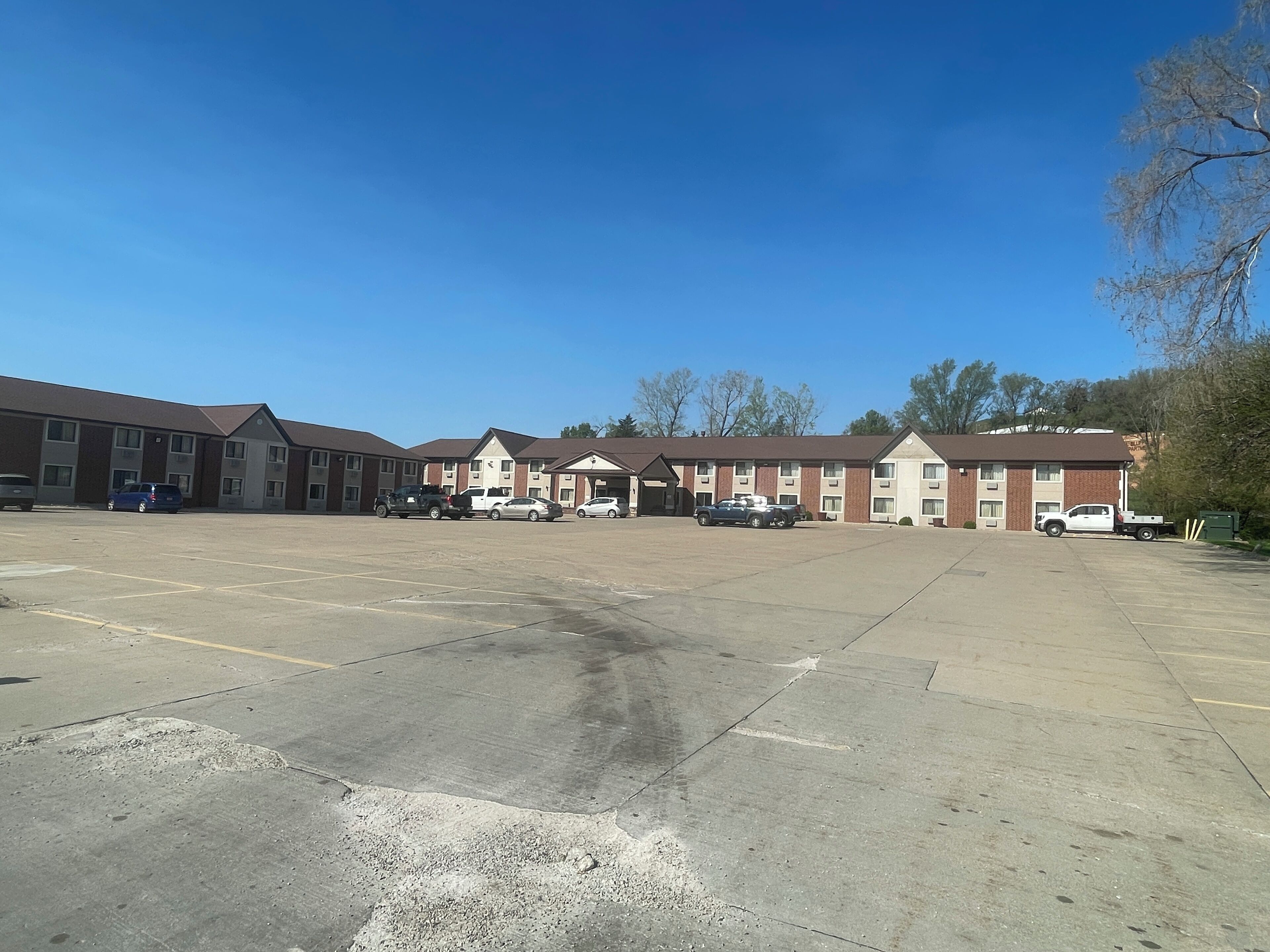 A parking lot with several vehicles, a motel building with multiple rooms, and a clear blue sky.