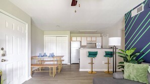 A modern living room with a grey sofa, a dining area with a wooden table and benches, and a kitchen with a white fridge and bar stools. - King Queen 2BR /walk to NRG /1mi to MD Anderson (Houston)