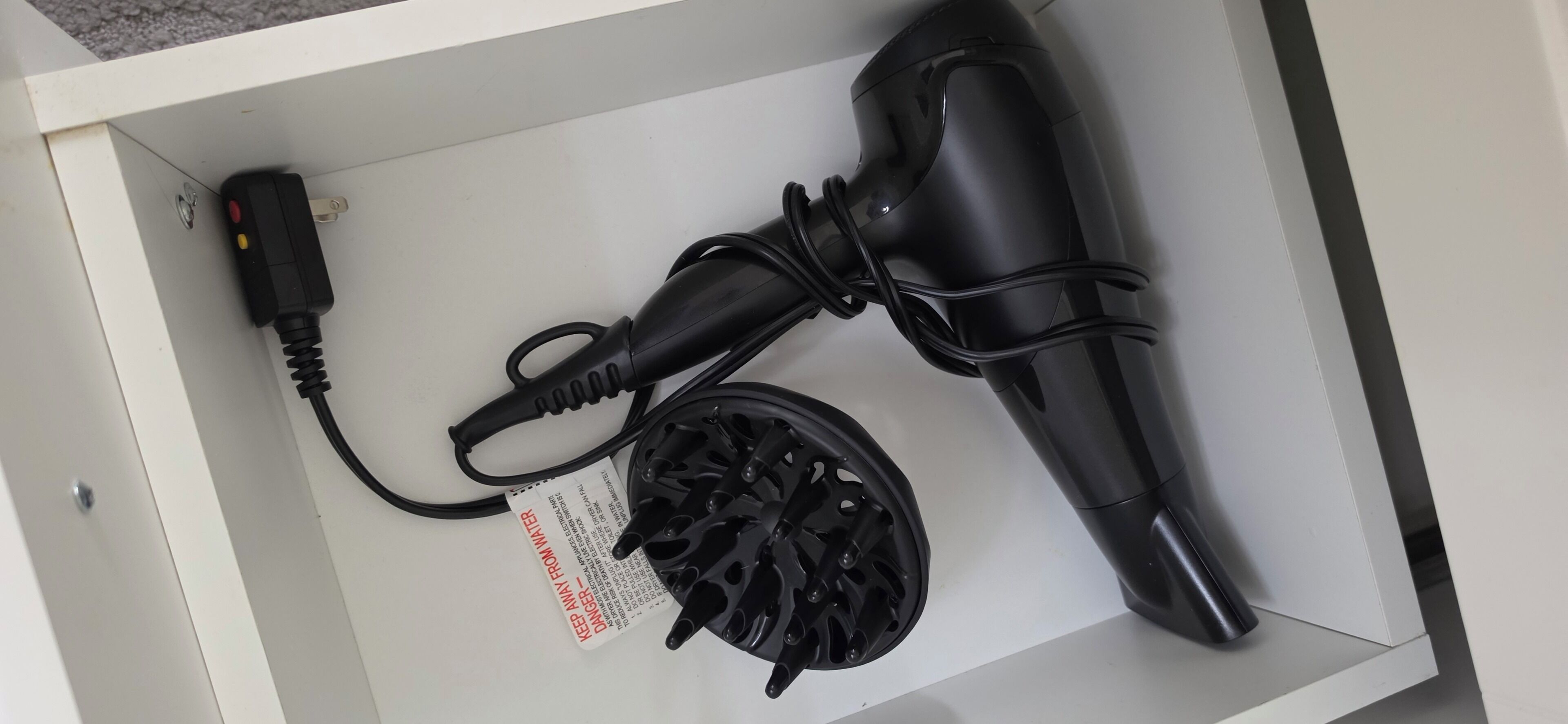 Un tiroir blanc contenant un sèche-cheveux noir, un câble d’alimentation noir et une étiquette d’avertissement de sécurité.