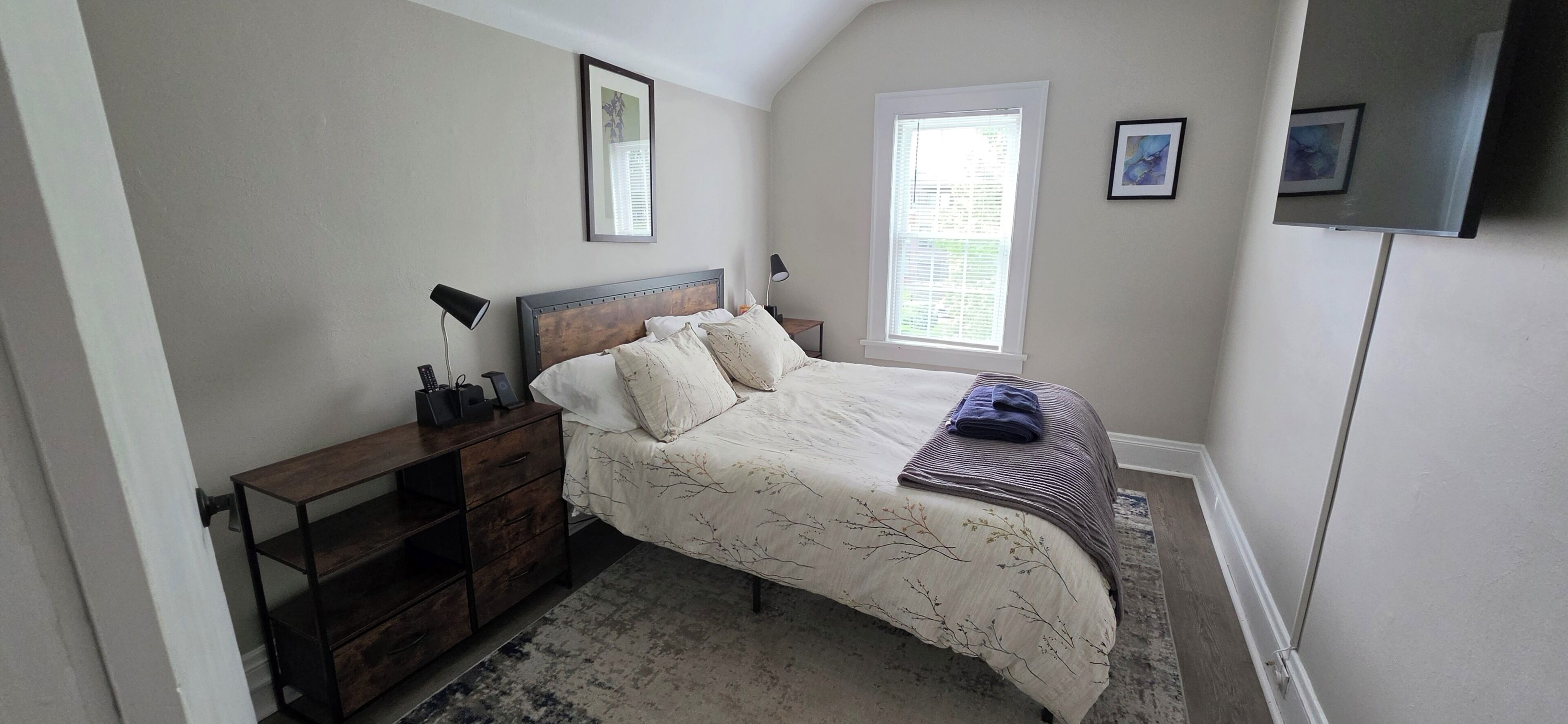 A bedroom with a bed, a nightstand, a dresser, a window, and a TV mounted on the wall.