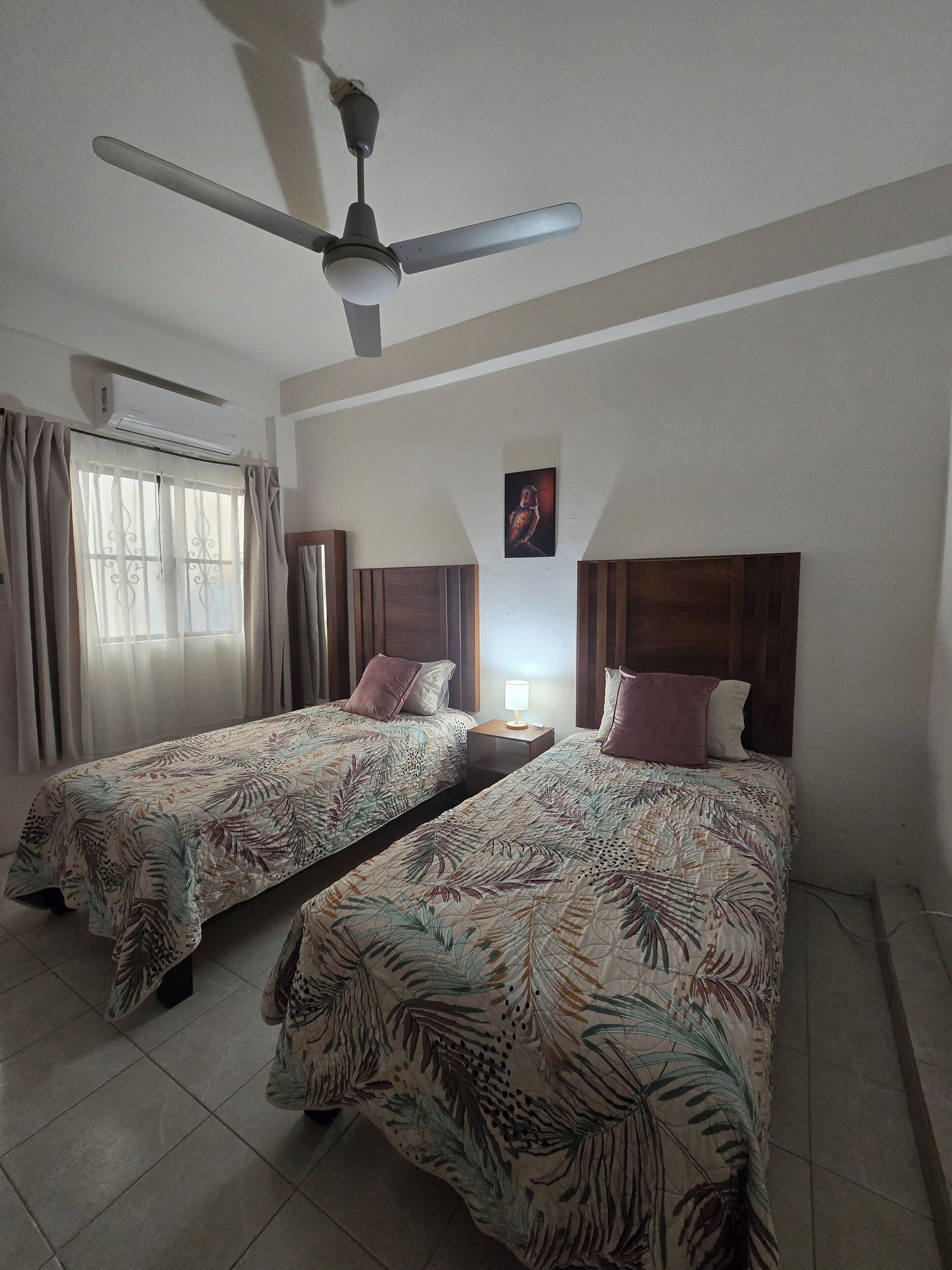 A hotel room with two beds, a ceiling fan, an air conditioning unit, a window with curtains, and a wall-mounted picture.