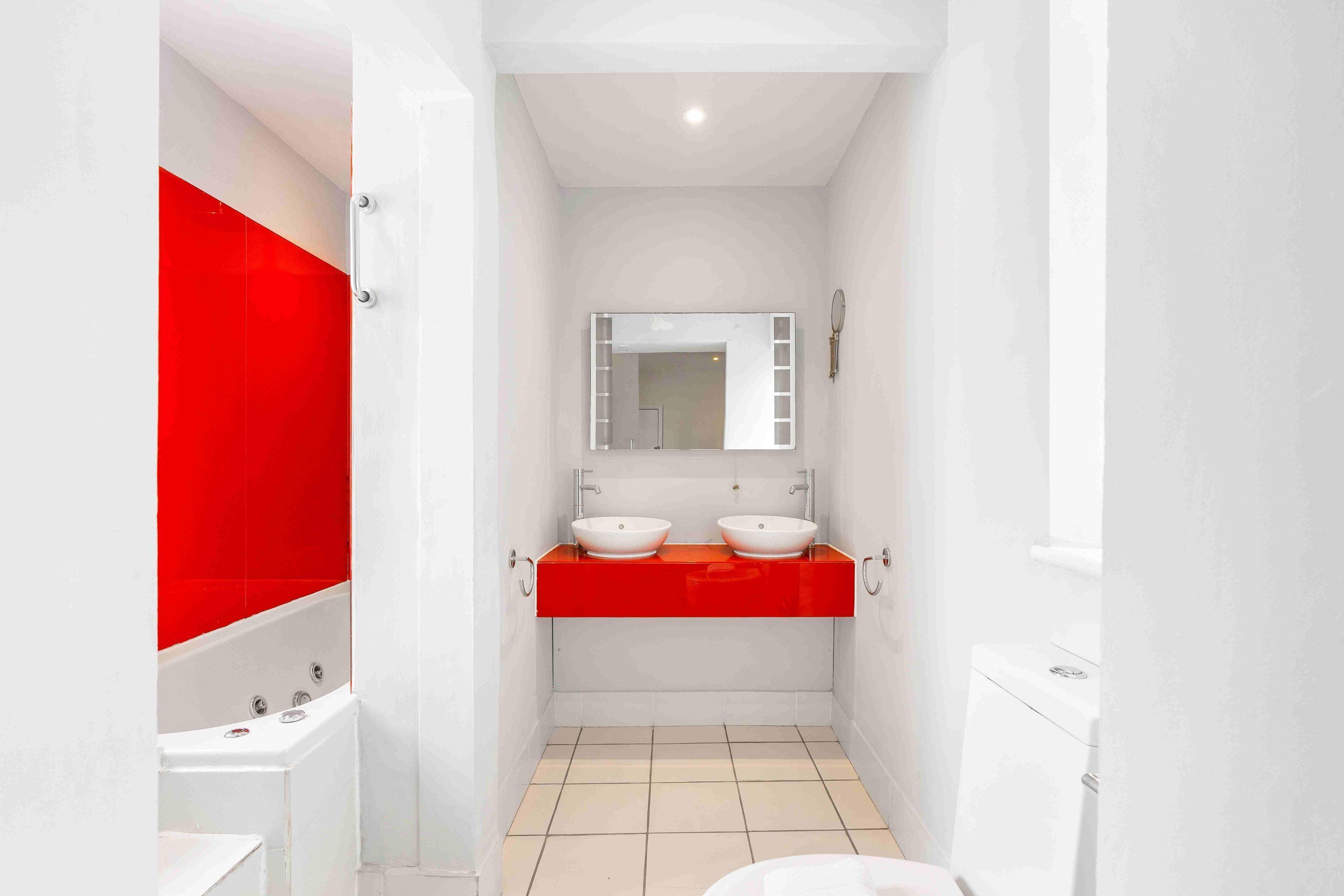 A bathroom with a white bathtub, two white sinks with red countertops, a mirror above the sinks, and a toilet.