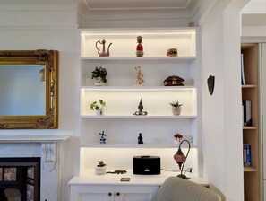 Ein Wohnzimmer mit Kamin, einem Spiegel mit aufwendiger Rahmenfassung und einer weißen Regaleinheit mit dekorativen Gegenständen.