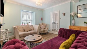 Ein Wohnzimmer mit einem beigen Sofa, einem runden Couchtisch, einem violetten Sofa, einem Holzkabinett und einer Kristallleuchter.