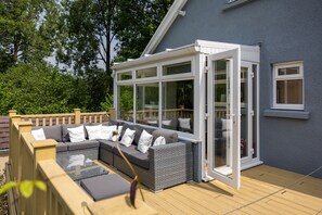A covered outdoor seating area with a glass enclosure, a wooden deck, and a grey building in the background. - Fern Hill - Close to Village and Beach (Saundersfoot)