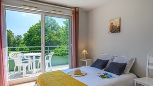 A bedroom with a bed, a nightstand, a lamp, a window with a view of greenery, and a painting on the wall. - À proximité de la plage | Accès piscine | Balcon (PLESCOP)