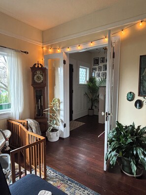 A room with a wooden crib, a large potted plant, a wall clock, and a doorway leading to another room with framed pictures. - Cozy East Van detached home with large backyard (Vancouver)