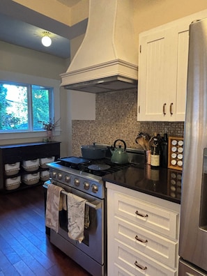 A kitchen with a stove, oven, white cabinets, and a window with a view of trees. - Cozy East Van detached home with large backyard (Vancouver)