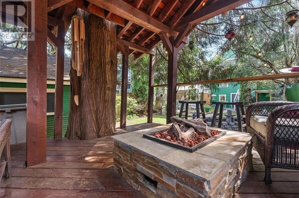 A covered outdoor patio with a stone fire feature, wicker furniture, and a large tree. - Cozy East Van detached home with large backyard (Vancouver)