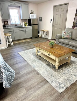 A modern kitchen with grey cabinets, a stainless steel refrigerator, and a microwave on top. There is a wooden coffee table with a plant, a grey sofa with a floral cushion, and a patterned rug on the floor.